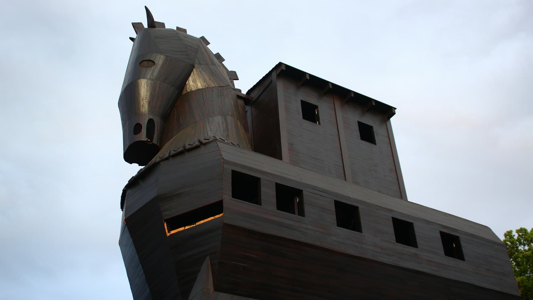A wooden Trojan horse with a hollow body and small windows, photographed against a pale blue sky.