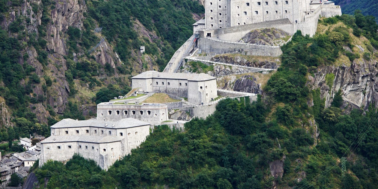 A stone hillside fortress of layered defensive walls and buildings climbing a steep, forested mountainside.