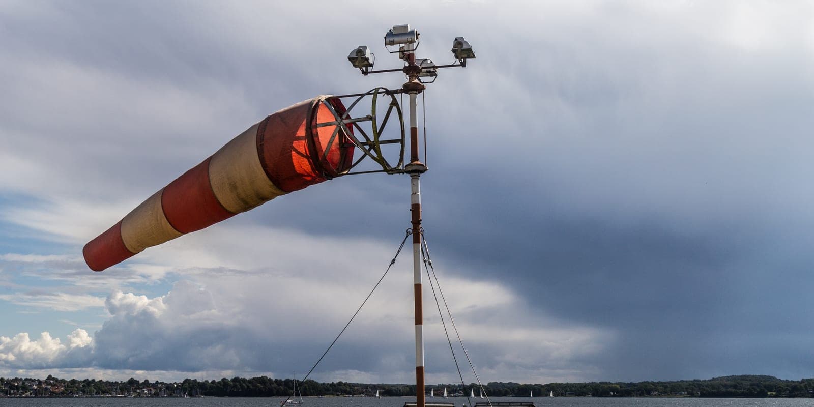 A red-and-white windsock stretched fully horizontal by strong wind on a harbour mast, under a moody overcast sky with sailboats on the water below.