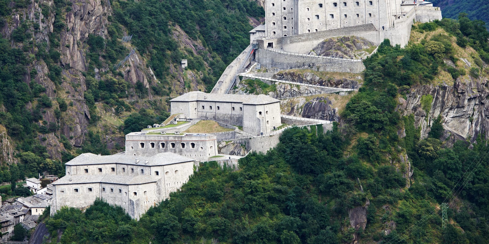 A stone hillside fortress of layered defensive walls and buildings climbing a steep, forested mountainside.